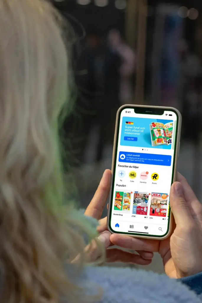 En person med blont hår håller i en smartphone som visar ett färgglatt gränssnitt för en shopping- eller kupongapp med olika erbjudanden och butikslogotyper. Telefonens skärm är ljus och tydlig, medan bakgrunden är oskarp.